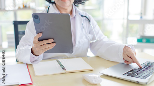 Asian smiling older woman doctor wearing medical coat stethoscope working reading data tablet laptop in healthcare office professional physician services hospital clinic wellness trust patient care.