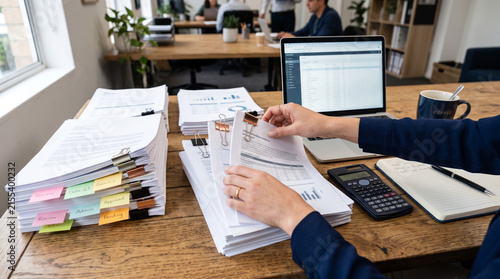 Busy office desk with stacks of documents, a laptop, a calculator, and a notebook. Hands are sorting through papers, indicating work in progress