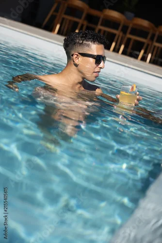 Fototapeta samoprzylepna Man is standing waist-deep in clear blue pool holding cocktail with sunglasses, deck chairs behind