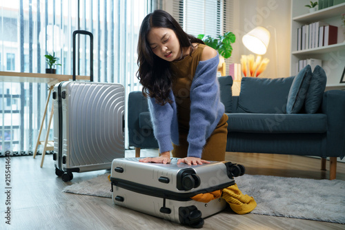 Young Asian Woman kneels on a rug Struggling to Close Overpacked Suitcase on Living Room Floor Before Trip, ideal for travel preparation and vacation planning content