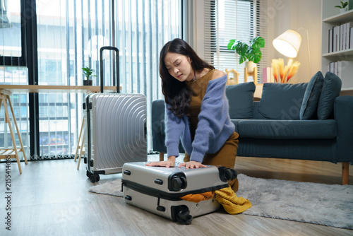 Young Asian Woman kneels on a rug Struggling to Close Overpacked Suitcase on Living Room Floor Before Trip, ideal for travel preparation and vacation planning content