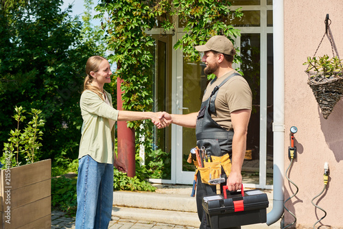 Woman shaking hands with male technician holding toolbox outside house. Image represents customer satisfaction and home repair services, suitable for local business marketing