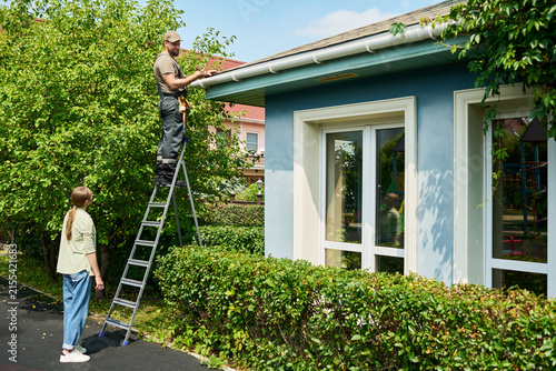 Caucasian male contractor standing on ladder repairing roof gutter while Caucasian female homeowner watches. Image represents home maintenance services and residential property care