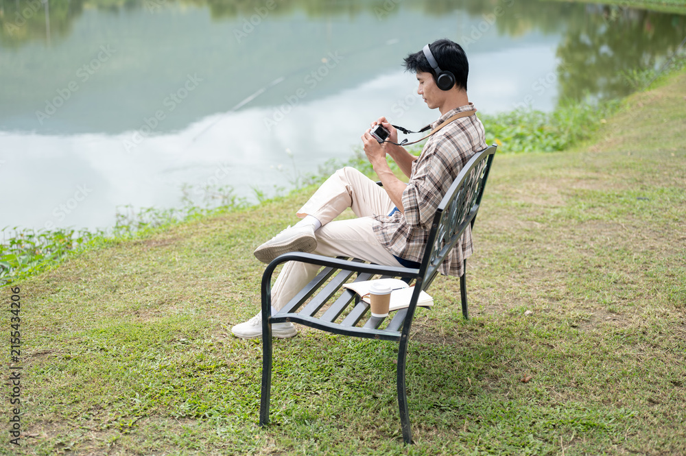 Fototapeta premium Asian man with headphones sitting alone on bench by lake and enjoy photographing the natural scenery