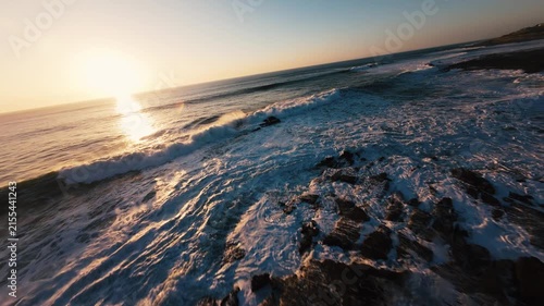 Sunset waves crash over dark rocks. Golden light reflects on foamy water. Sky transitions from blue to warm orange. Calm ocean meets rugged shore. Peaceful coastal scene. Great for travel, nature