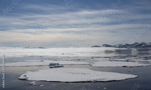 Broken sea ice in the Arctic Ocean off the coast of Svalbard with a calm sea, blue sky and white clouds. There is a low lying band of cloud obscuring the mountains on the shore.