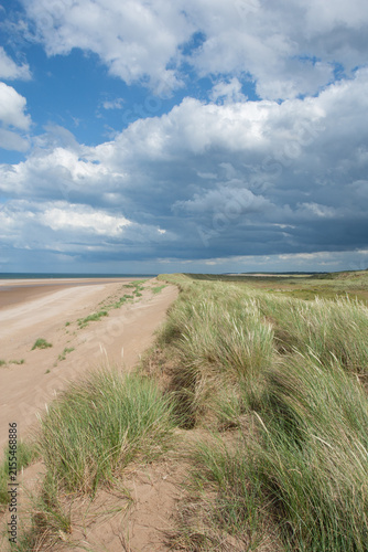 Sand dunes and marram grass on an English beach with blue skies and white clouds. The line of dunes provides a leading line into the photograph.