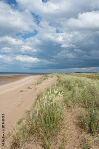 Sand dunes and marram grass on an English beach with blue skies and white clouds. The line of dunes provides a leading line into the photograph.