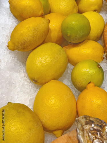 Fresh yellow lemons and a few green citrus fruits displayed on crushed ice at a market, showcasing vibrant colors and natural texture.
