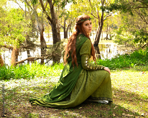 full length portrait of beautiful female model, kneeling pose figure, wearing green medieval historical gown costume with cape. Magical lake forest background with trees.

