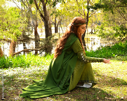 full length portrait of beautiful female model, kneeling pose figure, wearing green medieval historical gown costume with cape. Magical lake forest background with trees.
