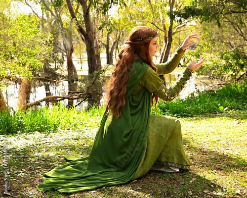 full length portrait of beautiful female model, kneeling pose figure, wearing green medieval historical gown costume with cape. Magical lake forest background with trees.
