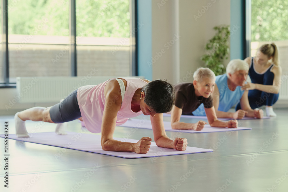 Fototapeta premium Mature Caucasian woman performing forearm plank on yoga mat during fitness class with senior people. Image represents active aging and is suitable for wellness marketing