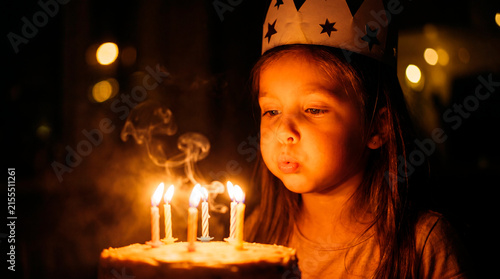 Little girl wearing paper crown blowing out candles on birthday cake in the dark. Warm cinematic candle lighting.