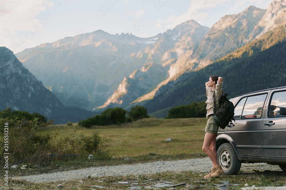Obraz premium A lone young woman with a backpack stands beside a parked car in a rugged mountain valley, preparing for a hike and enjoying a moment of solitude. A feeling of independence.