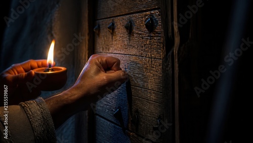 Hand knocking on a rustic wooden door beside a candle at night. Persistent prayer, seeking help, faith, and the biblical teaching ask and it will be given.