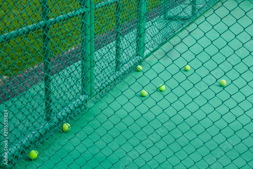 Tennis balls scattered on a green outdoor court viewed through a chain link fence