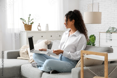 Pretty young African-American woman with laptop sitting on sofa at home