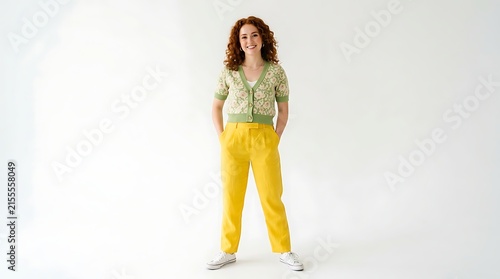 Full body portrait of a smiling young woman with curly red hair wearing a floral cardigan and yellow trousers isolated on white background