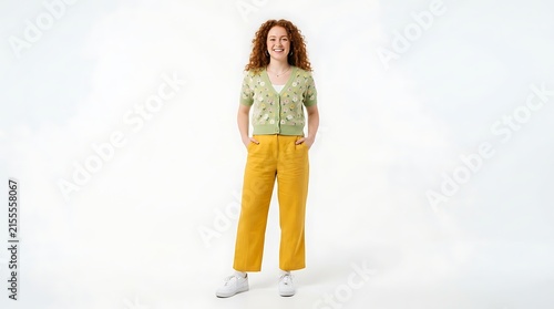 Full body portrait of a smiling young woman with curly red hair wearing a floral cardigan and yellow trousers isolated on white background
