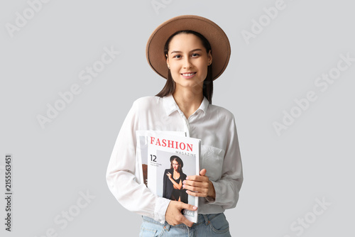 Beautiful young woman in stylish brown hat with magazine on grey background