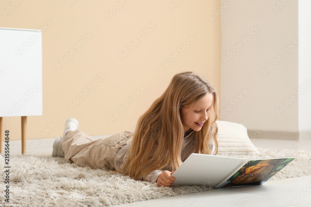Fototapeta premium Little girl reading book while lying on floor at home