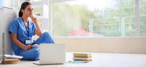 Фотография Thoughtful female medical student in blue scrubs and glasses studying on the flo