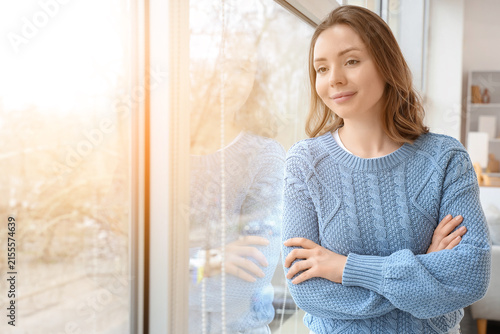 Young woman in a blue knitted sweater looking out a sunlit window with arms c...