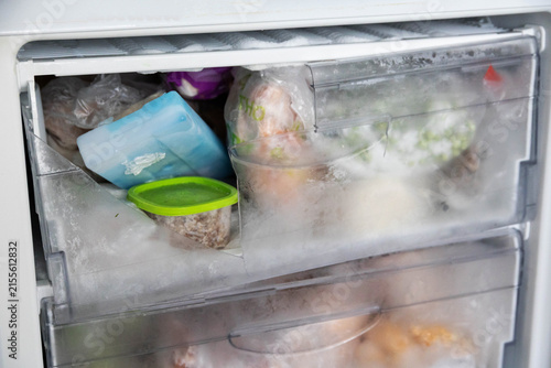 Broken plastic freezer drawer filled with frosted frozen food items and ice, showing damage and heavy ice accumulation inside an old refrigerator freezer compartment, requiring maintenance