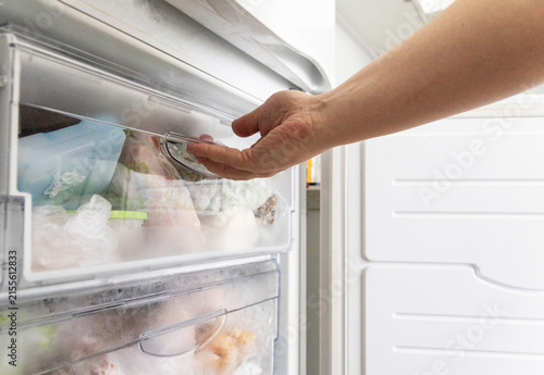 Human hand opening transparent drawer of modern white refrigerator freezer compartment filled with various frozen food items in plastic bags for storage and meal preparation