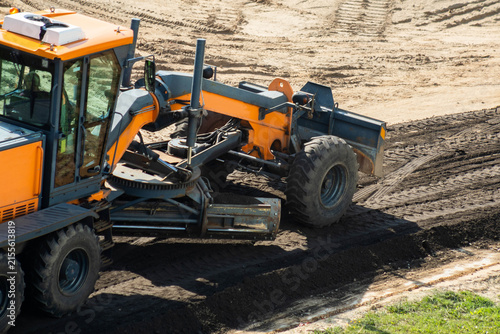 Large orange motor grader leveling roadbed surface, spreading and compacting asphalt chips on construction site, preparing for new road pavement during bright sunny day