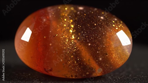 Close-up video of a sparkling orange gemstone with golden glitter inclusions, rotating on a dark background