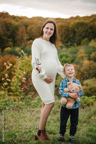 Happy mom and child. Pregnant woman and her little son holding a teddy bear outdoors in autumn. Smiling expecting mother standing with child on a scenic hill.
