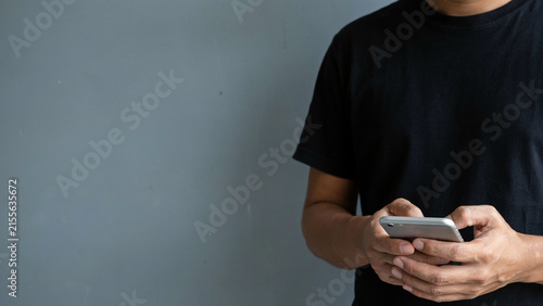 Young man in black t-shirt standing against a gray wall while using his smartphone to text and browse
