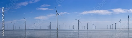 Panoramic view of Wind Farm Fryslân in the IJsselmeer, showing rows of offshore wind turbines reflected in calm water under a blue sky with soft clouds.