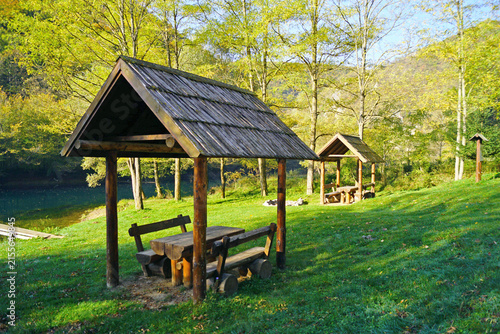 Canvas Print Wooden shelters with pitched shingle roofs on green lawn, autumn forest and river visible beyond