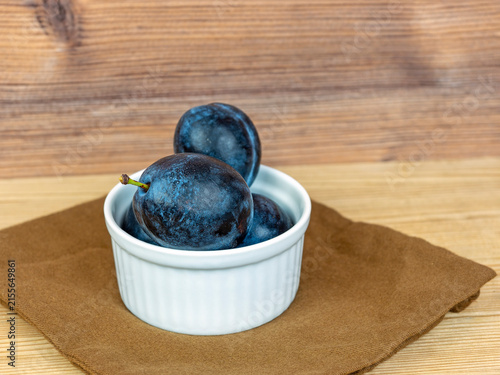 фотографія Rustic still life captures a white ramekin brimming with four ripe plums upon a square of rich, dark brown linen