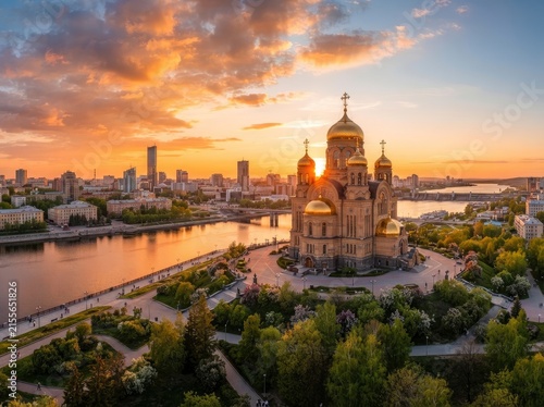 Aerial view of an orthodox cathedral with golden domes situated on a riverbank in a city park at sunset, surrounded by lush green trees and modern urban skyline in the background.