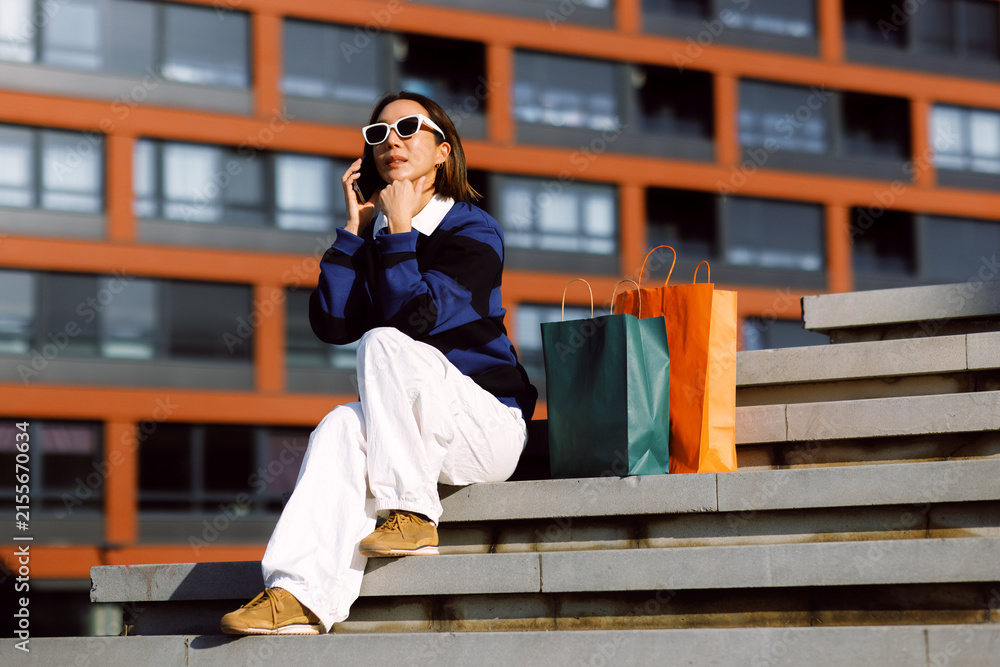 Fototapeta premium Young woman wearing sunglasses sitting on urban stairs talking on her smartphone with colorful shopping bags beside her