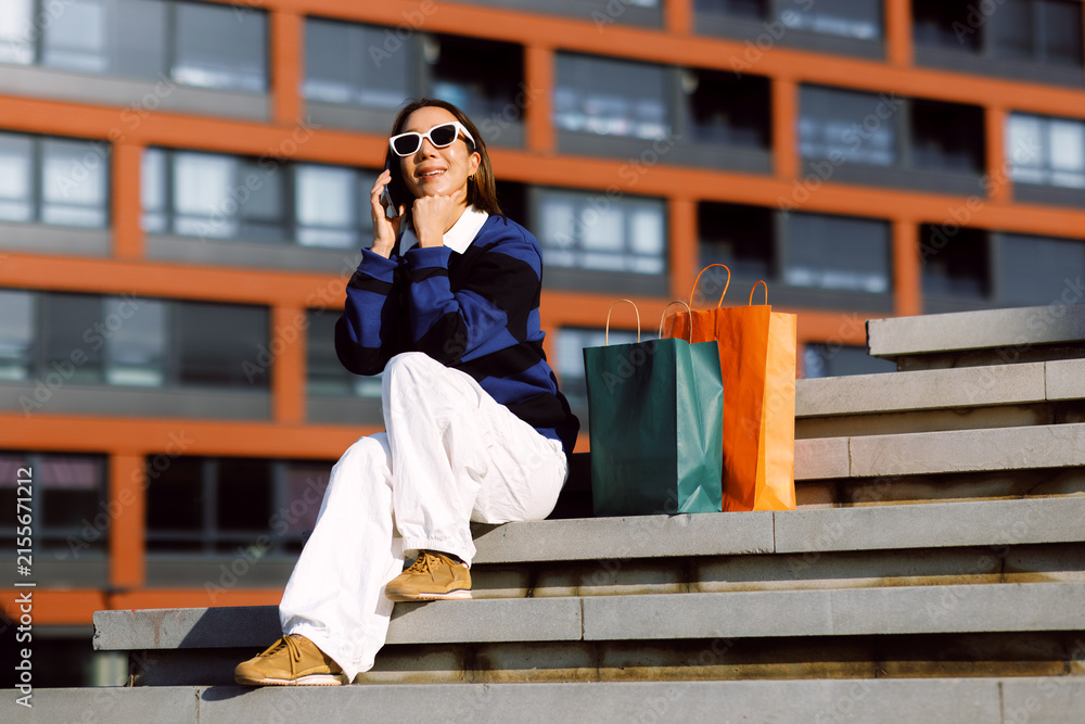 Fototapeta premium Smiling woman sitting on city steps resting with shopping bags while having a cheerful mobile phone conversation