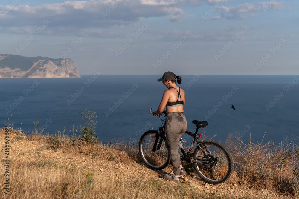 Fototapeta premium Bicycle travel woman standing on a cliff overlooking the sea while resting during an outdoor workout session in summer