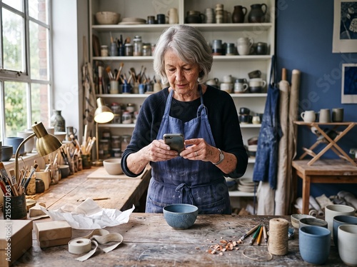 An older woman with gray hair, wearing an apron, is using her smartphone while standing at a workbench in a pottery studio. Shelves filled with pottery supplies are visible in the background
