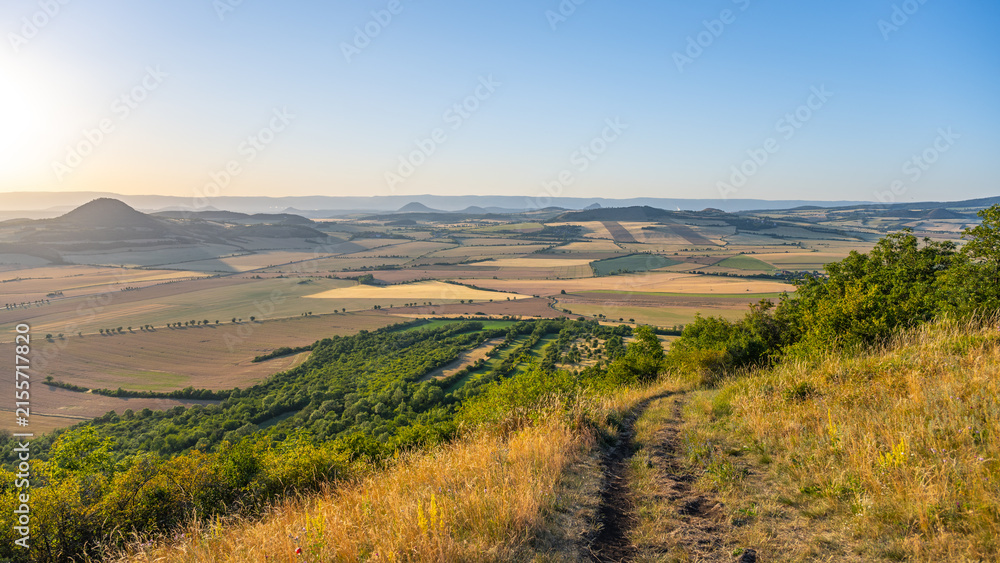 Obraz premium People watch the sunset from Oblik Hill. The sun sets over the fields and valleys in Czechia. The scenery shows rural land with trees and hills under a clear sky.
