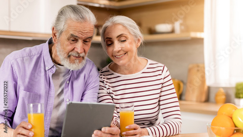 Принти на полотні Smiling Elderly Caucasian Man And Woman Sitting Together In Kitchen, Engaged With Tablet And Enjoying Fresh Orange Juice