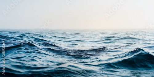 Deep blue ocean waves rolling gently across the vast horizontal sea surface under a bright hazy horizon during a calm and peaceful early morning sunlight session, minimal seascape