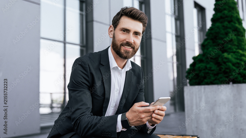 Fototapeta premium Smiling Caucasian Male Businessman In Formal Attire Holding Smartphone While Sitting On Bench At Modern Office Setting Engaging In Communication. Professional Lifestyle, Business Concept.