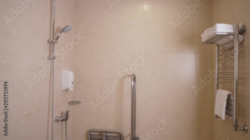 Clean modern bathroom featuring beige marble tiles, chrome shower head, grab bars, towel warmer with white towels. Soft lighting creates calm inviting mood