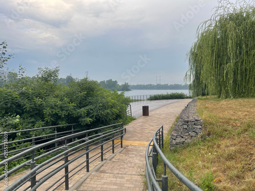 Scenic paved promenade along the waterfront park with weeping willow tree, metal handrails, and a view of the river under a stormy sky.