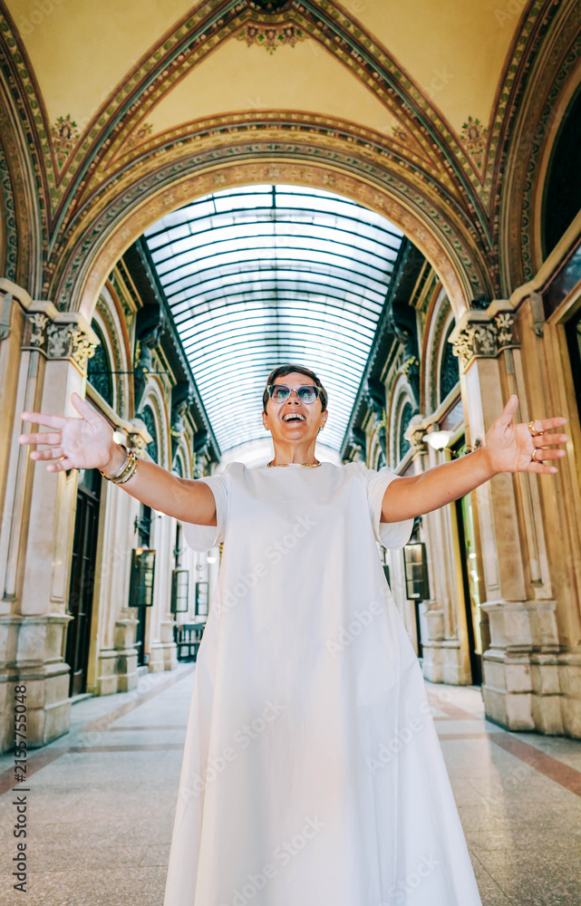 Obraz premium Woman in white dress with sunglasses stands in grand architectural corridor, arms outstretched, showcasing ornate arches and glass ceiling in a historic building