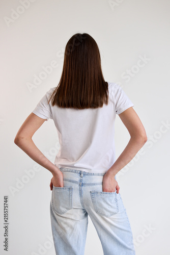 Rear view young woman standing with hands in back pockets, wearing white T shirt and light blue jeans against plain white studio background, showing casual posture, clean styling, minimal fashion